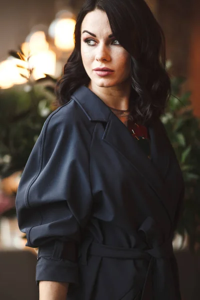 Estilo de vida y citylife. Hermosa dama de negocios morena en joyas modernas y abrigo marino de moda almorzando en la cafetería. Mujer se sienta junto a la mesa y sostiene la taza de té — Foto de Stock