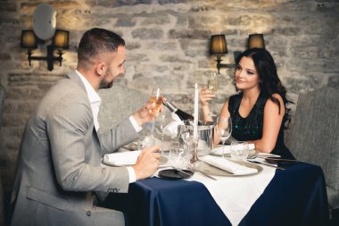 Romantizm ve flört konsepti. Şampanya bardaklı genç bir çift restoranda kutlama yapıyor ve kadeh kaldırıyor. İki kadın ve erkek şık bir gece elbisesi ve romantik bir akşam yemeği.