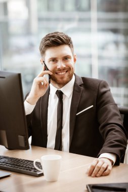 İş konsepti. Ofis masasında oturan genç işadamı cep telefonuyla yaptığı iş hakkında iyi haberler almaktan memnun. Camdan arka planda takım elbiseli bir adam.
