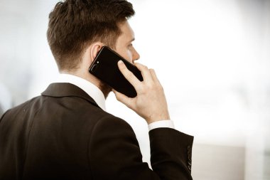 İş konsepti. Ofisteki genç işadamı elinde telefonla konuşmakla meşgul. Çok ciddi bir iş problemini çözüyor. Camdan arka planda takım elbiseli bir adam.