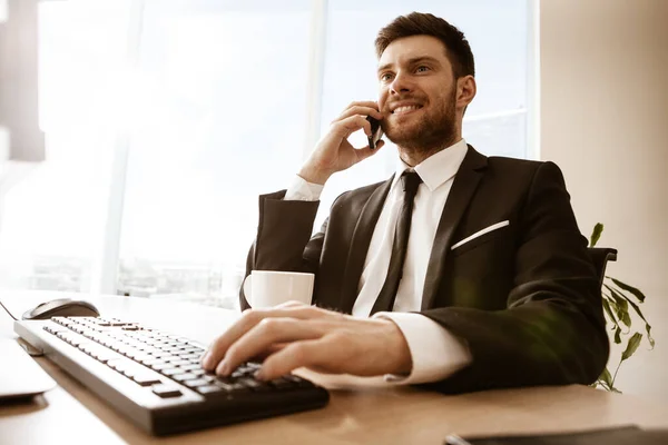 Conceito de negócio. Jovem empresário sentado na mesa do escritório feliz falando em um telefone celular recebendo boas notícias sobre seu trabalho. Mãos no teclado. Homem de terno dentro de casa no fundo da janela de vidro — Fotografia de Stock