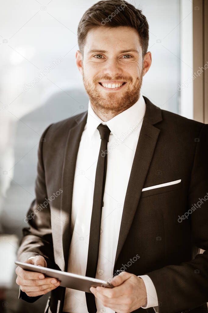 Business concept. Happy smiling young businessman standing in office holding wireless touchpad tablet computer. Man in suit indoors using internet device on glass window background