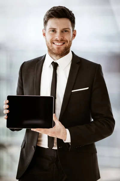Conceito de negócio. Feliz sorridente jovem empresário de pé no escritório demonstrando computador tablet touchpad sem fio. Homem de terno dentro de casa usando dispositivo de internet no fundo da janela de vidro — Fotografia de Stock