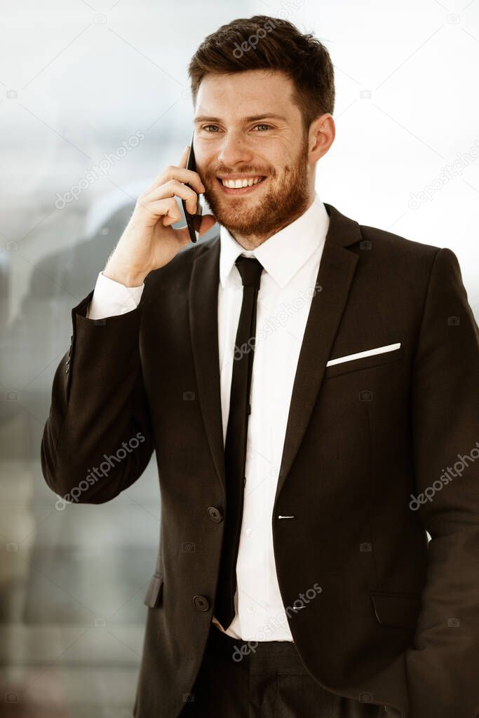 Business concept. Happy smiling young businessman standing in office talking on a cell phone getting good news about his work. Man in suit indoors on glass window background.