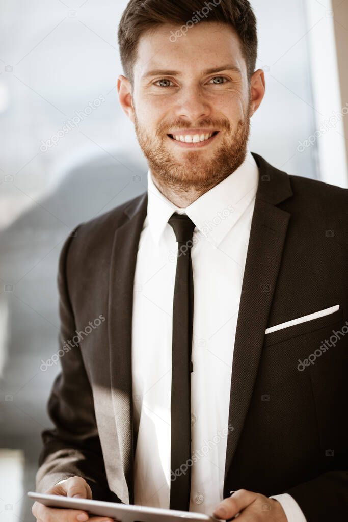 Business concept. Happy smiling young businessman standing in office holding wireless touchpad tablet computer. Man in suit indoors using internet device on glass window background.