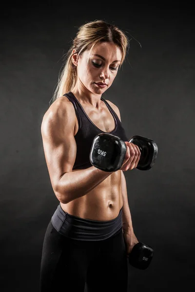 运动和健康的生活方式理念 美丽的女运动员在黑暗的背景下展示她受过训练的身体 健美女子模特儿用哑铃训练二头肌 — 图库照片