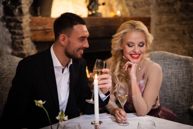 Romantizm ve flört konsepti. Şampanya bardaklı genç bir çift restoranda kutlama yapıyor ve kadeh kaldırıyor. İki kadın ve erkek şık bir gece elbisesi ve romantik bir akşam yemeği..