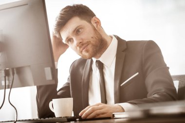 İş konsepti. Başarılı genç iş adamı iş başında. Ofis masasında oturan ve bilgisayarla çalışan bir yönetici. Pencere arkasında takım elbiseli meşgul bir adam..