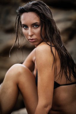 Yaz tatili konsepti. Formda, zayıf vücutlu, siyah mayo bikinili, taş üstünde kumsalda oturan genç ve seksi bir kadın. Moda kadın modeli deniz kenarında poz veriyor.
