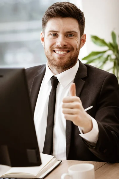 Concetto aziendale. Giovane uomo d'affari di successo al lavoro. Manager seduto al tavolo dell'ufficio felice mostrando pollice in su. Uomo sorridente in tuta all'interno su sfondo finestra di vetro — Foto Stock