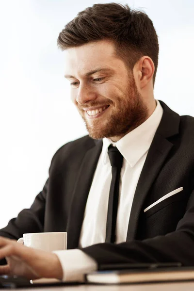 İşletme konsepti. Mutlu gülümseyen genç iş adamı ofiste oturup bilgisayarla çalışıyor. Camdan arka planda takım elbiseli bir adam. — Stok fotoğraf