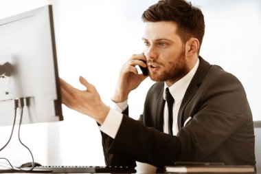 İş krizi konsepti. Ofis masasında oturan genç işadamı cep telefonuyla çok ciddi bir iş problemini çözmekle meşgul. Camdan arka planda takım elbiseli bir adam..