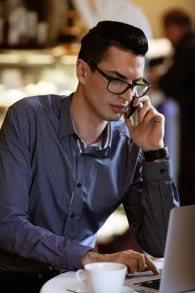 LGBTQ community lifestyle concept. Young homosexual man sits at the table in old-fashioned city cafe. Handsome gay male businessman talks on a phone while having a break at lunch.