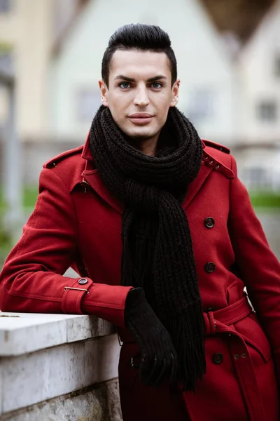LGBTQ community lifestyle concept. Young homosexual man stands near stone fence. Handsome fashionable gay male model poses in cityscape outdoors. Wears red coat, gloves, and black scarf.