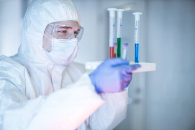 Corona virüsü konsepti. Maskeli, gözlüklü ve koruyucu giysili erkek bilim adamı salgın sırasında COVID-19 test tüpünü mavi sıvıyla inceliyor. Biyokimya laboratuvarındaki adam..