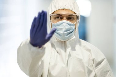 Corona virüsü konsepti. Maskeli, gözlüklü ve koruyucu elbiseli bir bilim adamı COVID salgını sırasında elinde lateks eldivenle dur işareti gösteriyor. Biyokimya laboratuvarındaki adam..
