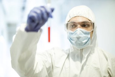 Corona virüsü konsepti. Maskeli, gözlüklü ve koruyucu giysili erkek bilim adamı salgın sırasında COVID-19 test tüpünü mavi sıvıyla inceliyor. Biyokimya laboratuvarındaki adam..