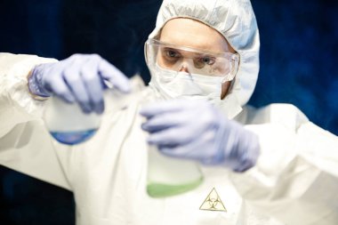 Corona virüsü konsepti. Maskeli, gözlüklü ve koruyucu elbiseli erkek bilim adamı COVID salgını sırasında laboratuvarda renkli sıvılarla dolu cam mataralar kullanarak biyokimyasal deney yapıyor..