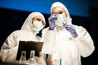 Corona virüsü konsepti. Erkek ve kadın mikrobiyoloji uzmanı maskeli, gözlüklü ve koruyucu elbiseli doktorlar test tüpünü incelerken COVID-19 salgını karantinasında analiz ediyorlar.