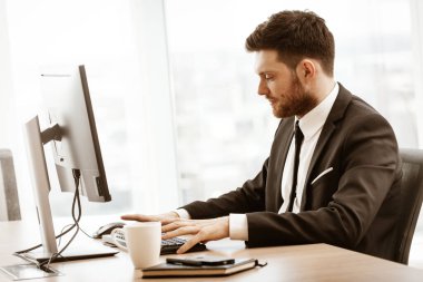 İş konsepti. Başarılı genç iş adamı iş başında. Ofis masasında oturan ve bilgisayarla çalışan bir yönetici. Pencere arkasında takım elbiseli meşgul bir adam..