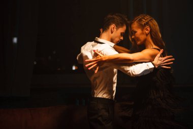 Dans ve aşk konsepti. Zarif gece elbiseli genç bir çift dramatik ışıkla dolu odada poz veriyor. İki dansçı kadın ve erkek birbirlerine tutkulu bir şekilde sarılırlar..