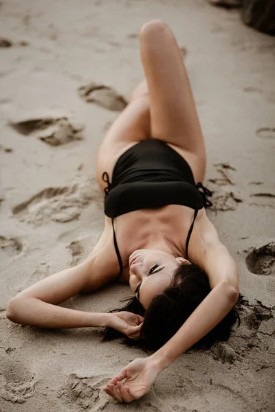 Koncepcja rekreacji letniej. Piękna młoda seksowna kobieta z wytrenowanym szczupłym ciałem w czarnym bikini leży na plaży. Moda kobieta model stwarza nad morzem — Zdjęcie stockowe