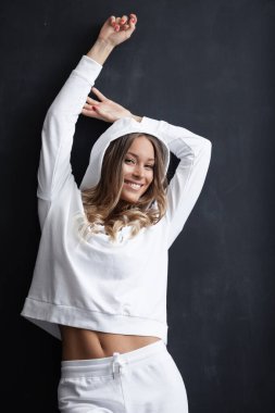 Sportif yaşam tarzı konsepti. Beyaz takım elbiseli, gülümseyen güzel bir kadının portresi. Seksi kadın model kapüşonlu ve pantolonu karanlık stüdyo arka planında.