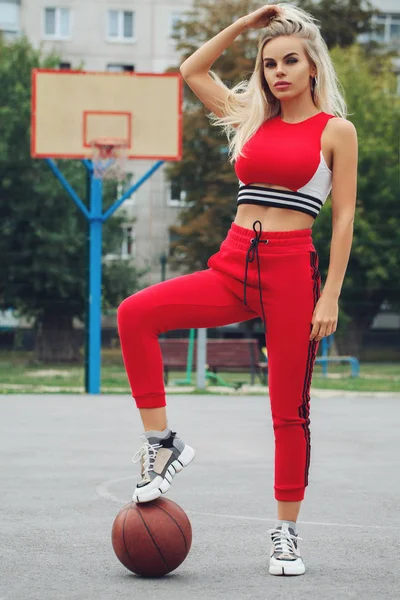 Beautiful sexy fitness blonde girl in red sport wear with perfect body with basket ball at basketball court. Sport, fitness, lifestyle concept