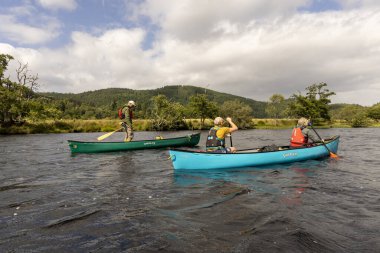İskoçya, Birleşik Krallık, 4 Ağustos 2025: Üç kişi bir gölde kanolarda. Kanolar mavi ve yeşildir. Gökyüzü bulutlu..