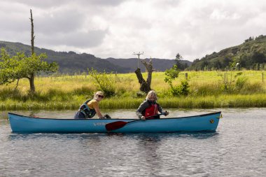 İskoçya, Birleşik Krallık, 4 Ağustos 2025: İki kadın sakin bir gölde mavi bir kanoda kürek çekiyor. Gökyüzü kapalı..
