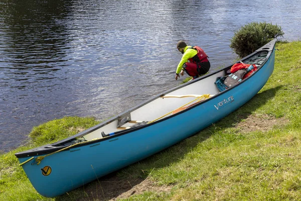 İskoçya, Birleşik Krallık, 4 Ağustos 2025: Bir adam mavi bir kanonun yanında diz çöküyor. Kano çimlerin üzerine park edilmiş, bir su kütlesinin yanında..