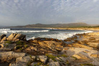 Küçük bir dalganın kayalara çarptığı kayalık bir kıyı şeridi. Gökyüzü bulutlu ve güneş gökyüzünde alçak.