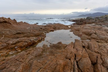 Ortasında küçük bir su kütlesi olan kayalık bir sahil. Su sakin ve gökyüzü bulutlu.