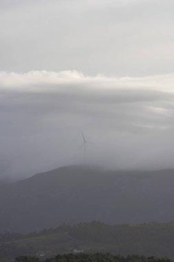 Bir dağ sırasının üstündeki gökyüzünde bir rüzgar türbini görülebilir. Gökyüzü bulutlu ve görüntünün havası biraz kasvetli.