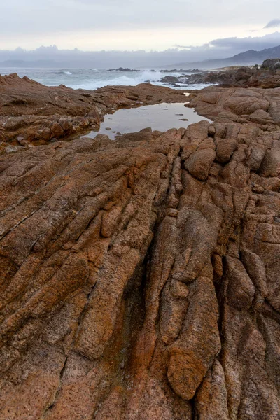 Arka planda büyük bir su kütlesi olan kayalık bir kıyı şeridi. Kayalar pürüzlü ve pürüzlü, ve su sakin görünüyor.