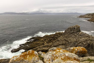Okyanus sakin ve gökyüzü bulutlu. Kıyıdaki kayalar yosunla kaplı.