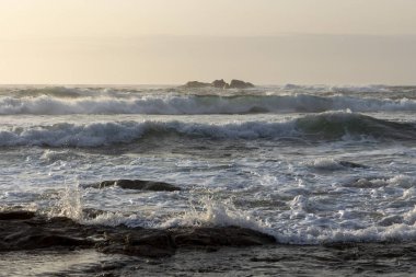 Okyanus sakin ve dalgalar küçük. Gökyüzü bulutlu ve güneş batıyor.