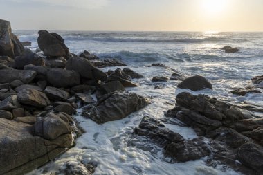 Okyanus sakin ve güneş batıyor. Kayalar kıyı şeridi boyunca dağılmış. Su beyaz ve gökyüzü mavi.