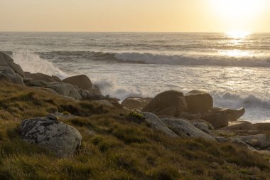 Okyanus sakin ve güneş batıyor. Kıyıdaki kayalar dağılmış ve çimenler kurumuş.