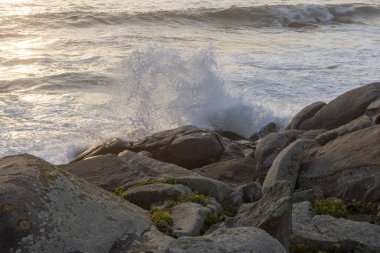Büyük bir dalganın kayalara çarptığı kayalık bir kıyı şeridi. Su beyaz, kayalar gri ve kahverengidir. Sahne, dalgaların sesiyle huzurlu ve huzurlu.