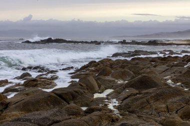 Okyanus engebeli ve kayalar büyük. Gökyüzü bulutlu ve su dalgalı.
