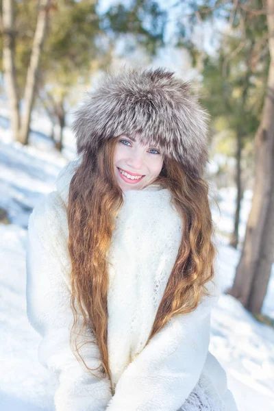 Girl in a white fur coat and a fur hat, with blue eyes and long hair against the background of snow and trees. Winter