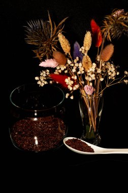 Kavanozdaki keten tohumlarının fotoğrafı ve siyah arka planda kurutulmuş çiçeklerle dolu bir kaşık.