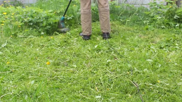 青いTシャツと医療用マスクの背の高い男が芝刈り機で芝生を刈る 暖かい夏の天気 草の風に移動します 民家の庭で雑草やタンポポの制御 — ストック動画