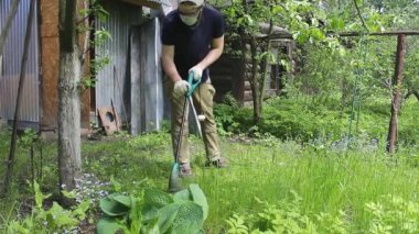 Mavi tişörtlü ve tıbbi maskeli uzun boylu bir adam elektrikli çim biçme makinesiyle çimleri biçiyor. Sıcak yaz havası, rüzgarda hareket eden çimenler. Özel bir evin bahçesinde ot ve karahindiba kontrolü..