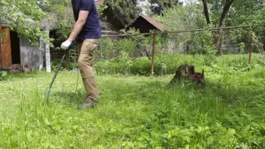 Mavi tişörtlü ve tıbbi maskeli uzun boylu bir adam elektrikli çim biçme makinesiyle çimleri biçiyor. Sıcak yaz havası, rüzgarda hareket eden çimenler. Özel bir evin bahçesinde ot ve karahindiba kontrolü..