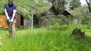 Mavi tişörtlü ve tıbbi maskeli uzun boylu bir adam elektrikli çim biçme makinesiyle çimleri biçiyor. Sıcak yaz havası, rüzgarda hareket eden çimenler. Özel bir evin bahçesinde ot ve karahindiba kontrolü..
