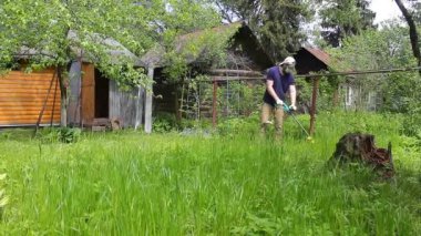 Mavi tişörtlü ve tıbbi maskeli uzun boylu bir adam elektrikli çim biçme makinesiyle çimleri biçiyor. Sıcak yaz havası, rüzgarda hareket eden çimenler. Özel bir evin bahçesinde ot ve karahindiba kontrolü..