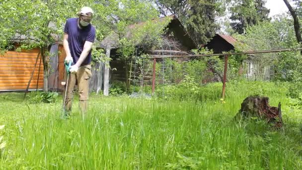 一个穿着蓝色T恤和医疗面罩的高个子男人用电动割草机修剪草坪 温暖的夏天天气 草地在风中飘扬 私家花园的杂草和蒲公英控制 — 图库视频影像