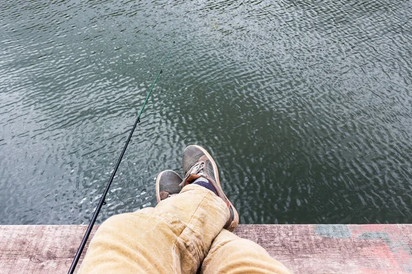 a man's legs hang from a wooden pier, and a fishing rod is lying nearby. recreation and fishing in nature.